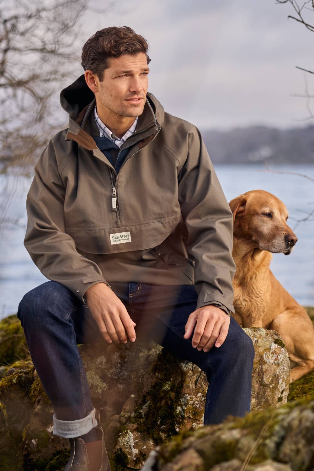 Man sitting on a rock with a dog by a lake wearing a brown jacket.