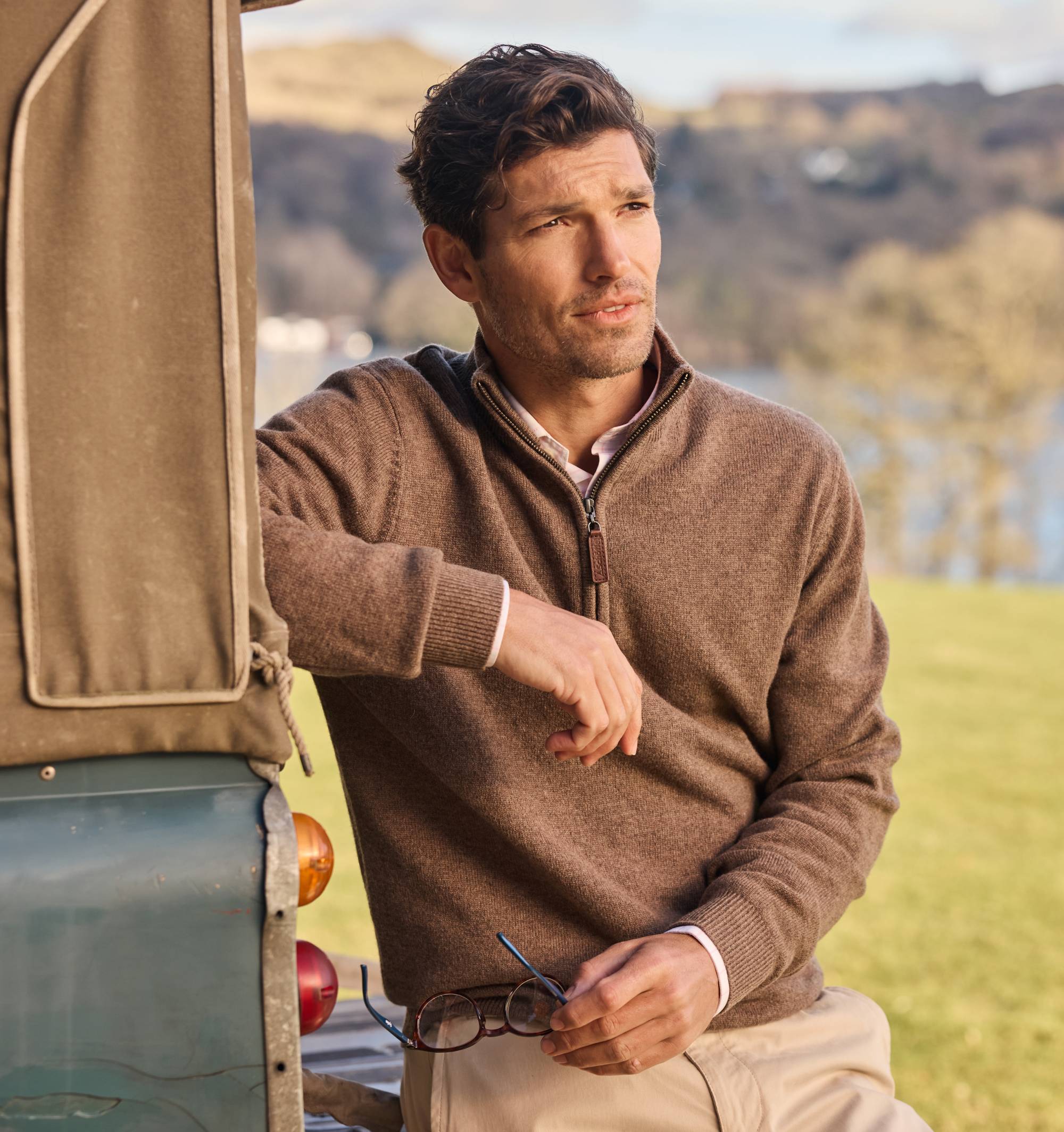 Man in a brown knitted sweatshirt standing next to a vehicle with a scenic background