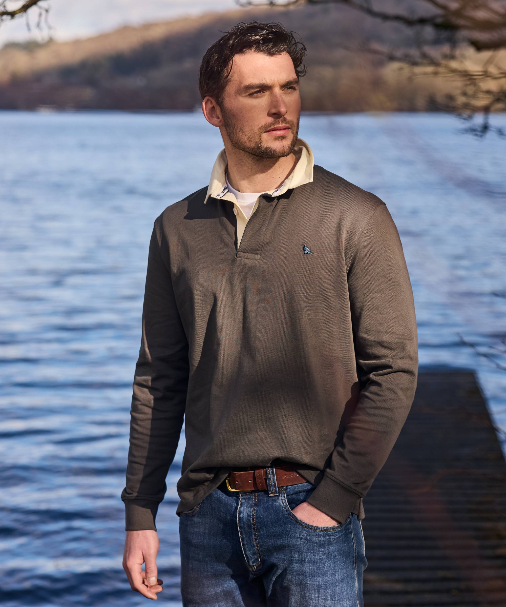 Man wearing a green long-sleeve rugby shirt and blue jeans standing by a lake.