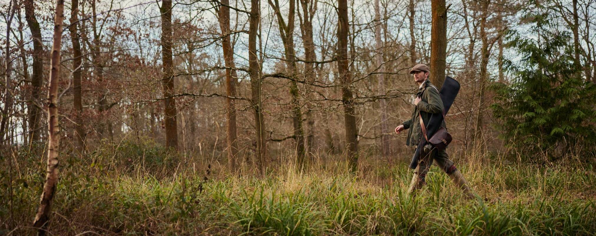 A man walking through woodland surrounded in trees and greenery. He is wearing countrywear and holding a leather bag on his shoulder.