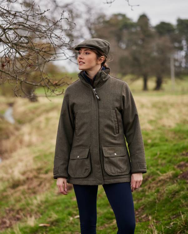 A woman wearing a tweed jacket, matching tweed cap and blue jeans, she is standing in a relaxed pose looking away from the camera.