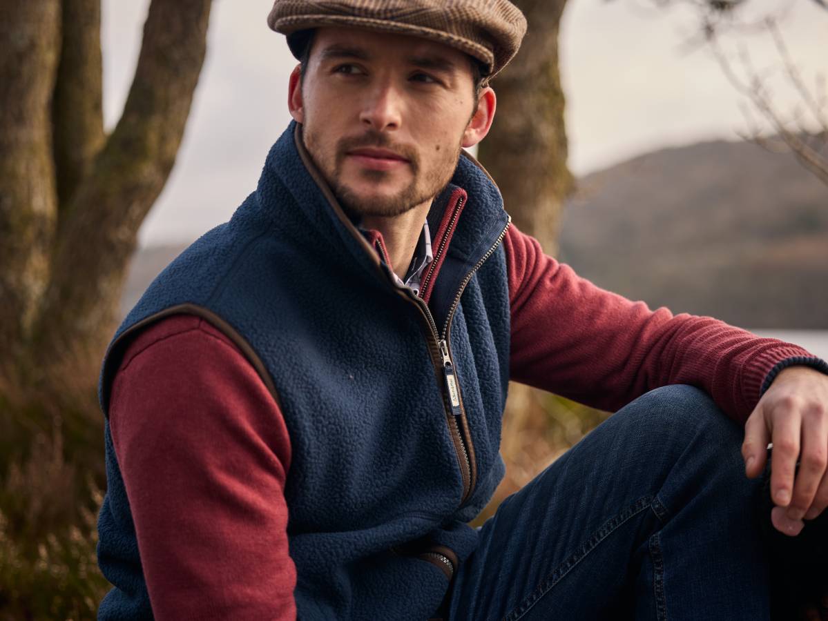Man wearing a blue gilet over a red sweatshirt with a cap, sitting outdoors.