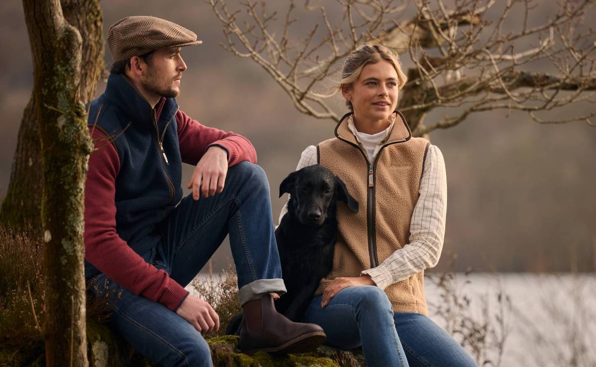 Two people sitting with a black dog by a lake, surrounded by trees and nature.