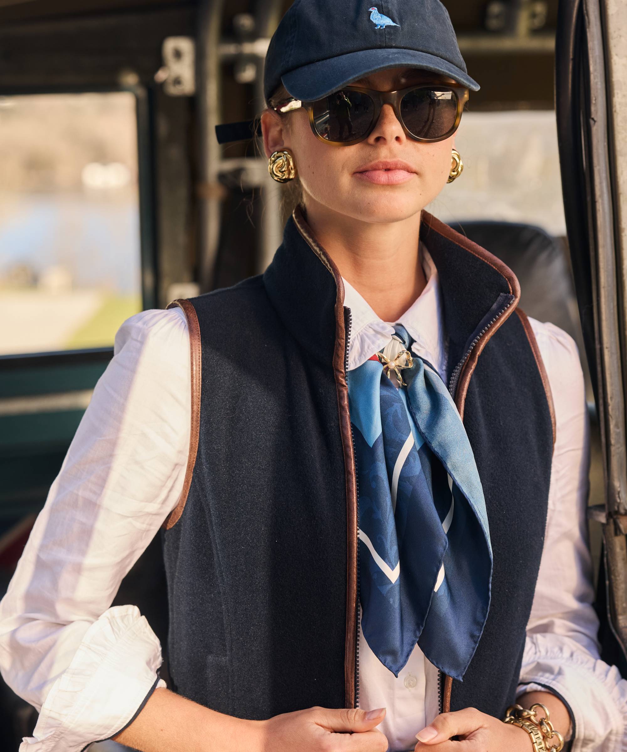Person wearing a navy cap, sunglasses, and a blue gilet with a white shirt and blue scarf.