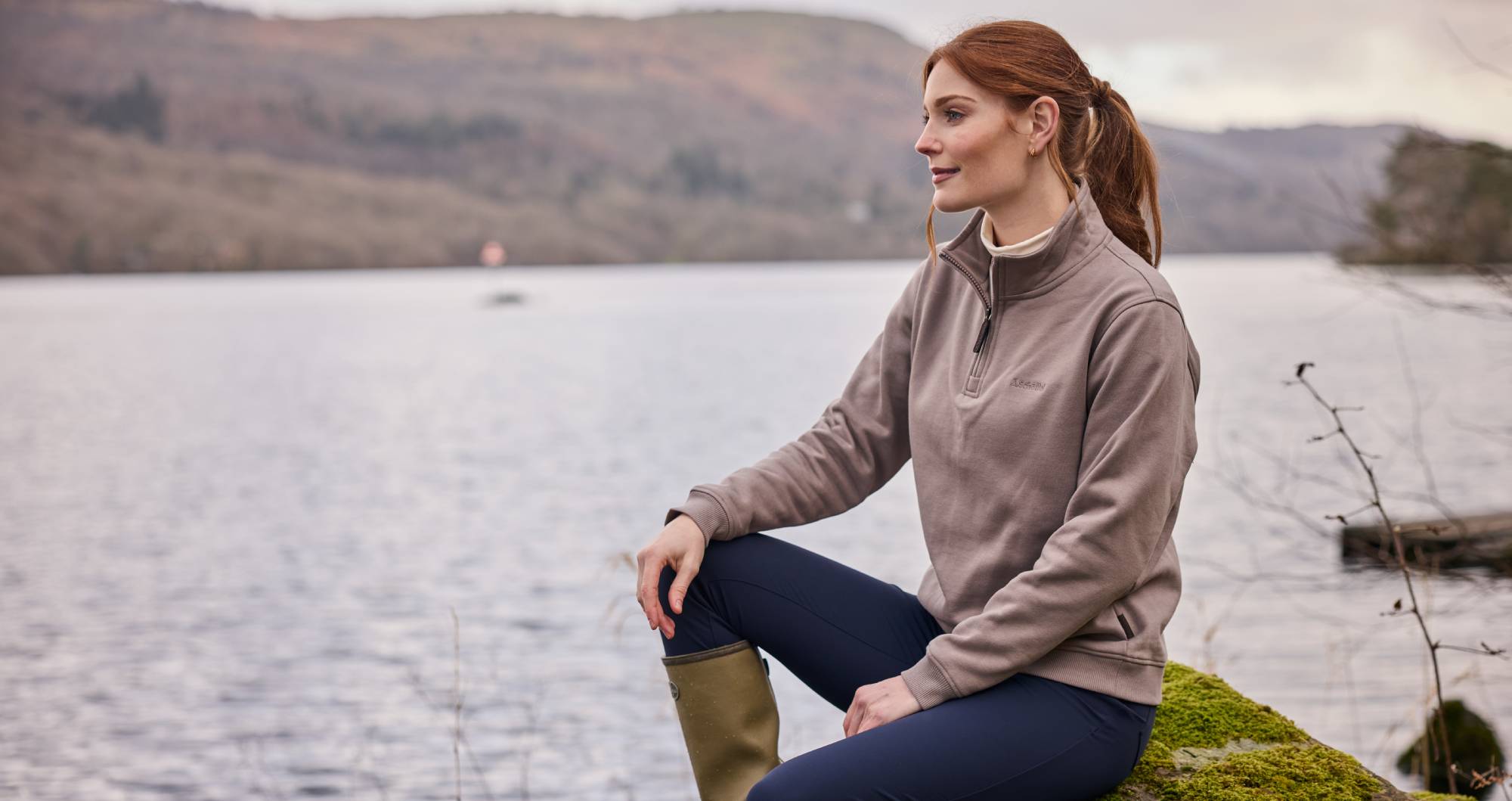 Woman sitting on a rock by a lake wearing a brown sweatshirt and navy trousers.
