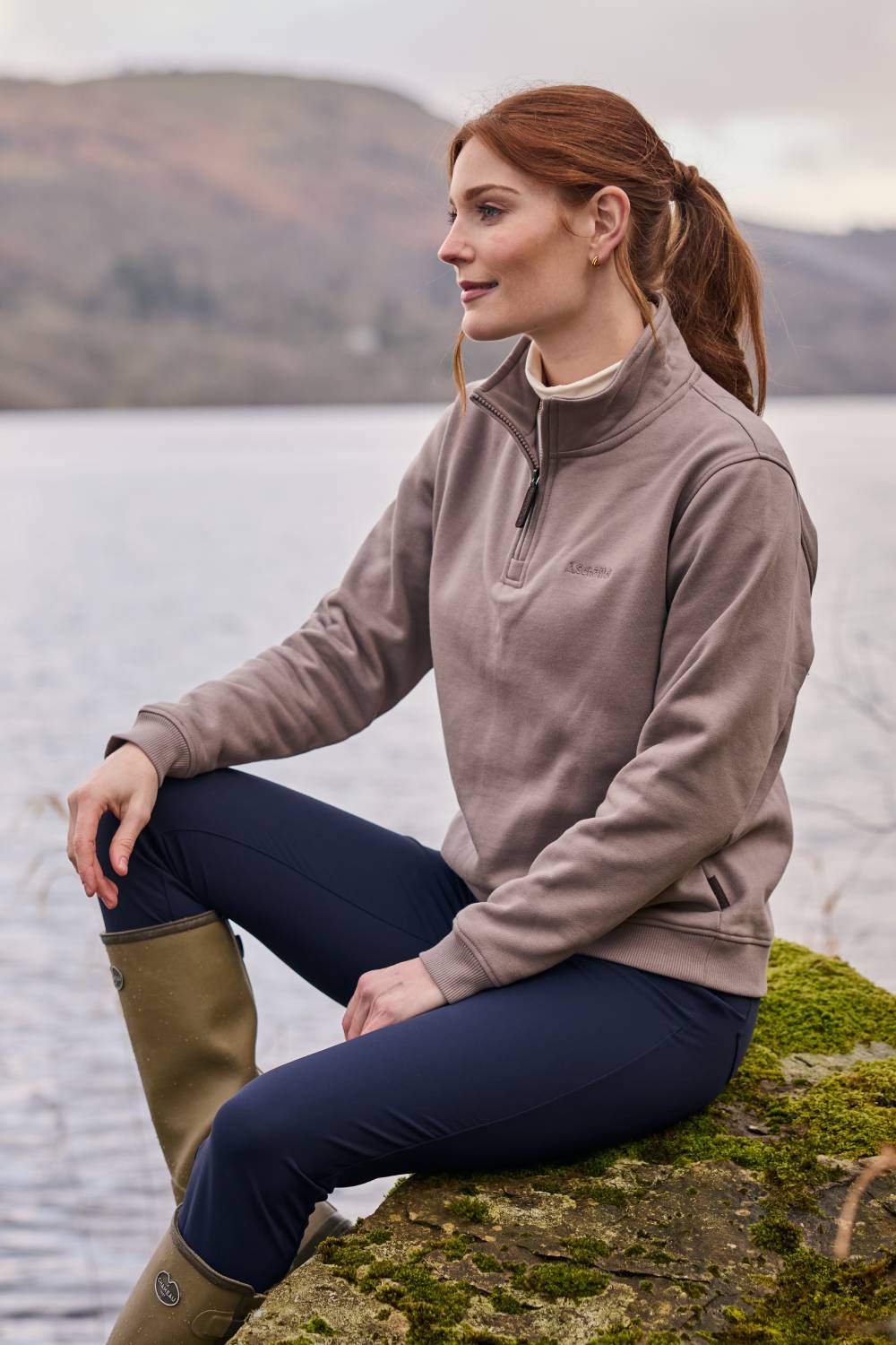 Woman sitting on a rock by a lake wearing a brown sweatshirt and navy trousers.