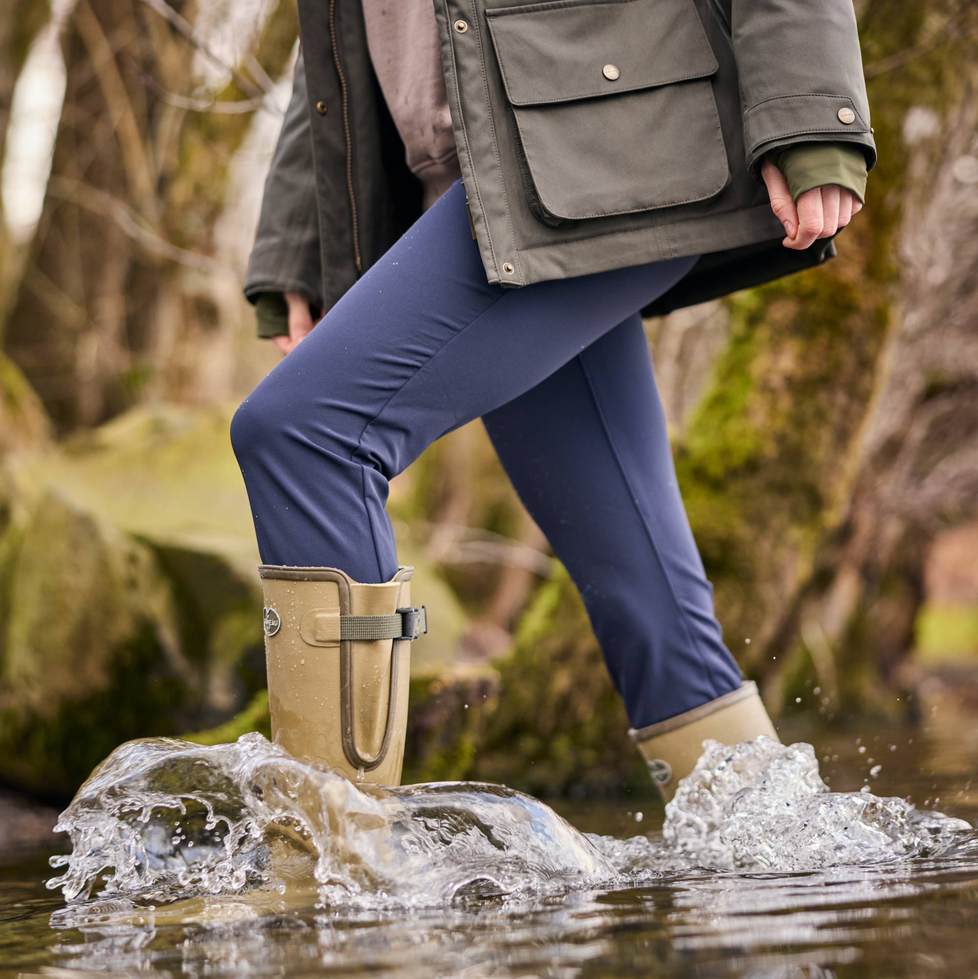 Person wearing blue trousers and green boots walking through water in a natural setting