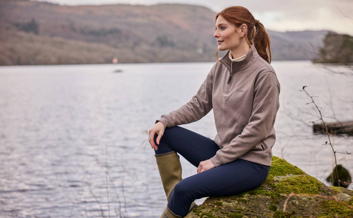 Woman sitting by a lake wearing a brown sweatshirt and dark blue trousers.