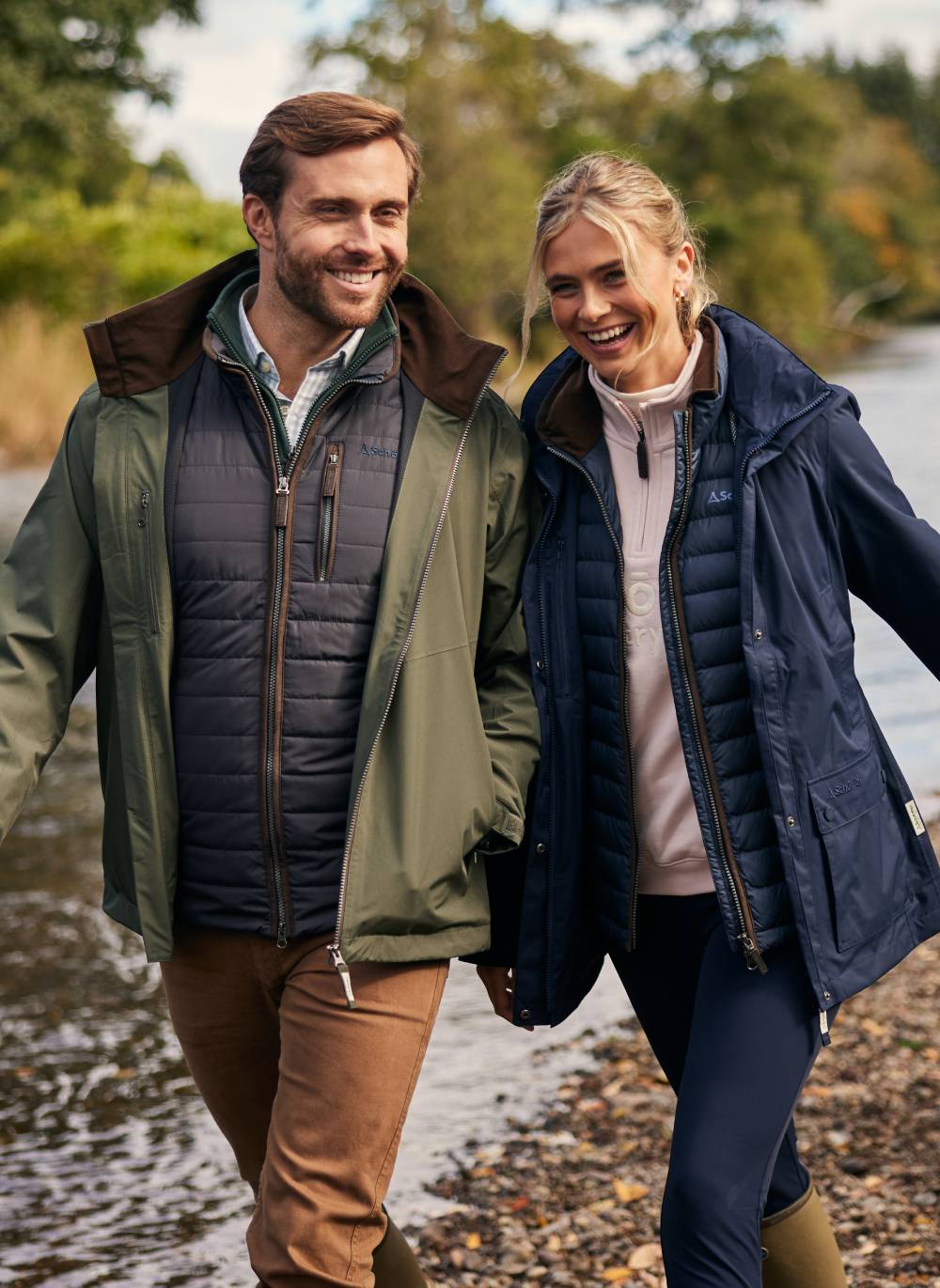 A smiling man and woman walk along the edge of a scenic river, dressed in stylish outdoor clothing. The man is wearing a green waterproof jacket over a navy quilted vest and a checkered shirt, while the woman wears a navy raincoat over a light-coloured sweatshirt. The background features lush green trees and a gently flowing river.