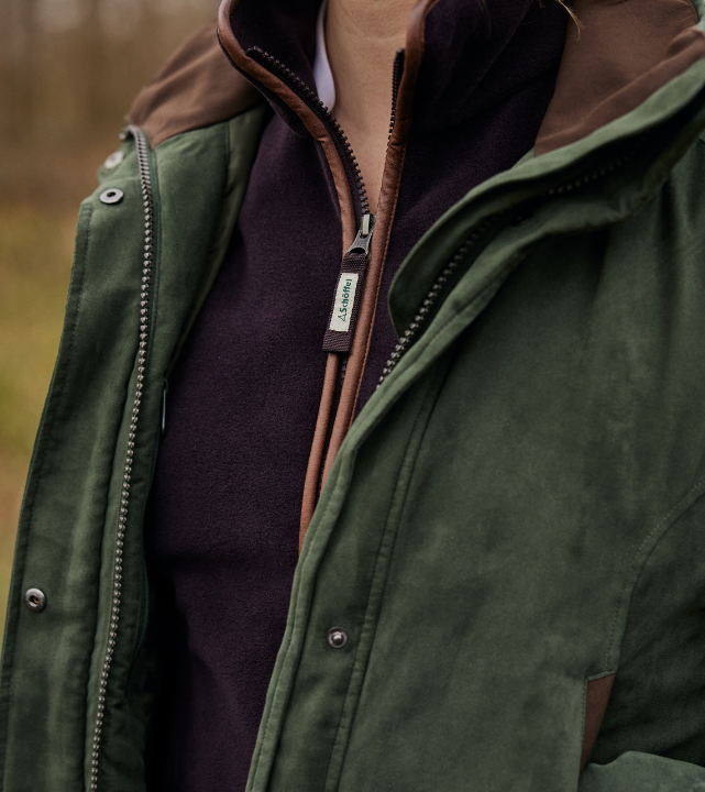 Close-up of a cedar green shooting coat layered over inner garments, showing zip, collar and contrast panel details.