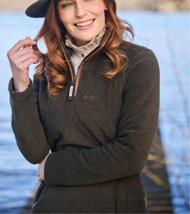 A smiling woman wearing a dark olive quarter-zip fleece with a brown leather-effect zip trim and a small embroidered Schöffel logo on the chest. A check shirt collar is visible underneath. She is wearing a dark cap and standing outdoors near water in bright sunlight.