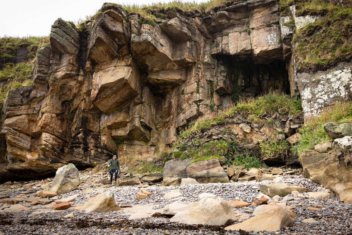 Rocky coastal cliff with grassy patches, pebble beach and person at its base.