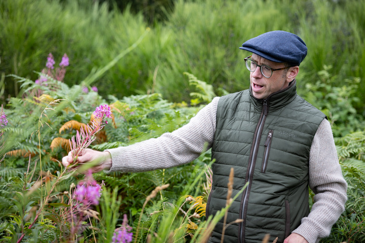 A man wearing a dark green quilted vest stands in a lush green garden, reaching toward pink wildflowers.