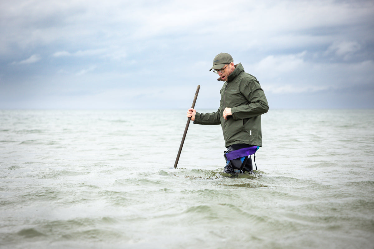 Man in an olive waterproof jacket and purple waders holds a wooden pole in shallow water.