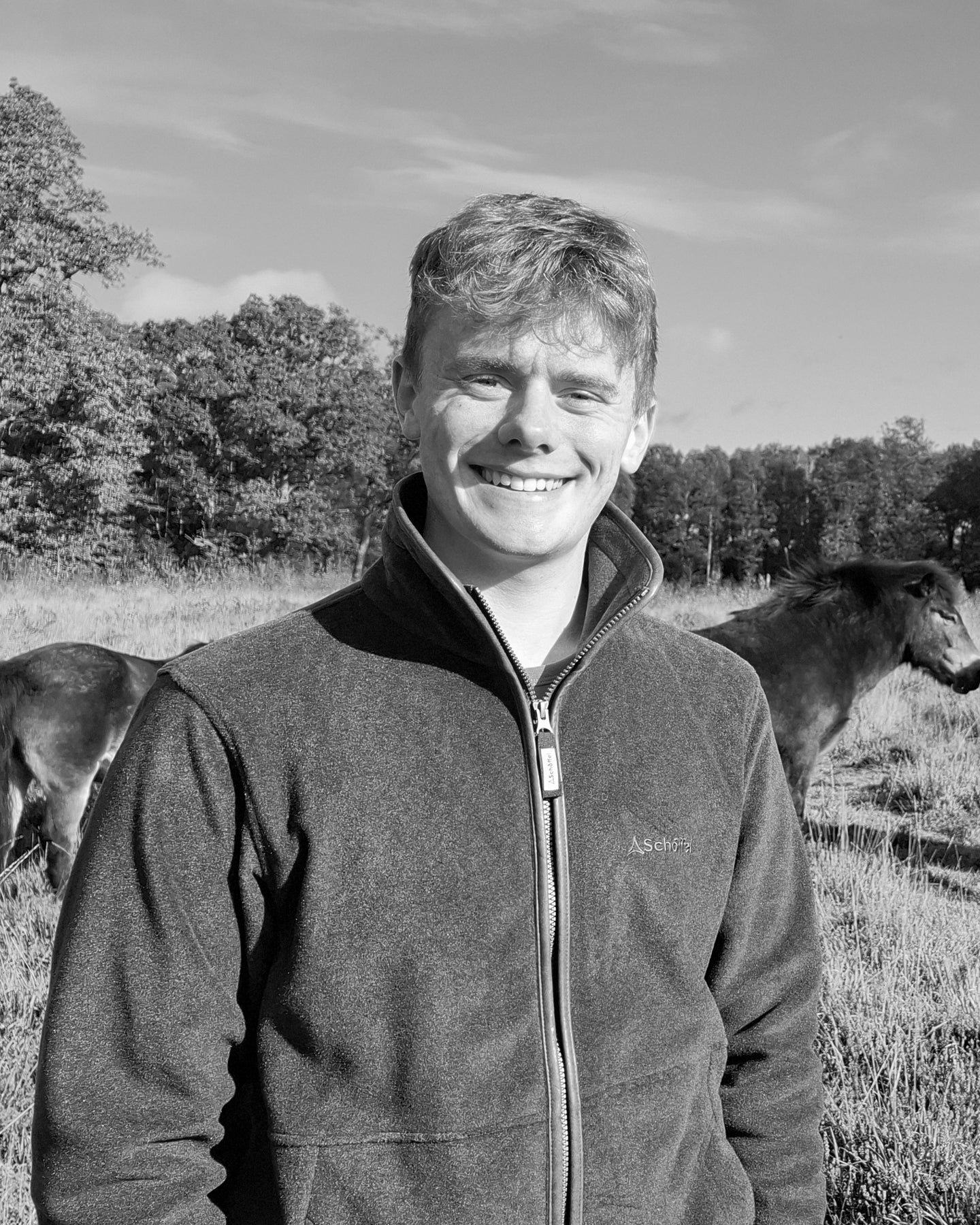 Tom Hilder standing in a field with horses, wearing a Schöffel jacket with a visible brand logo.