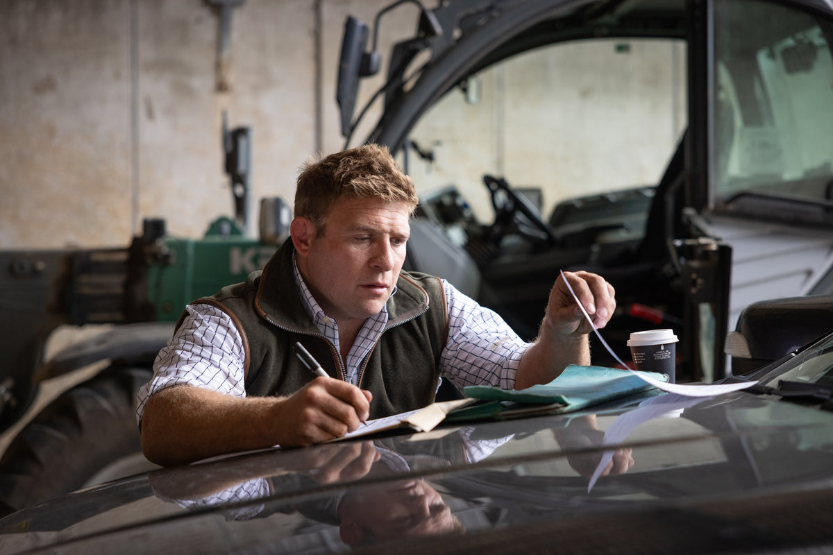 Tom Youngs in a gilet and check shirt doing paperwork on a tractor bonnet in the farm shed