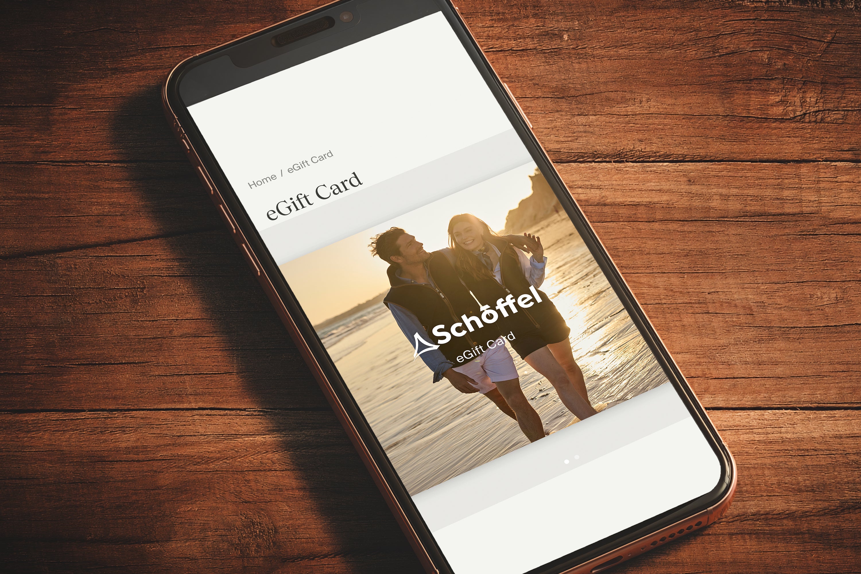 A smartphone lying on a dark wooden surface, displaying an eGift Card screen with the Schöffel logo and a photo of two people by the sea.