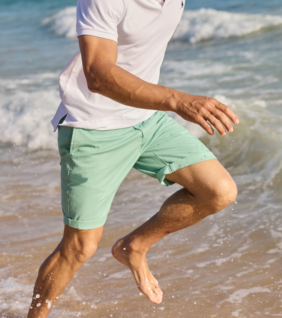 A man running through shallow ocean water on a beach, wearing a white polo shirt and mint green shorts.