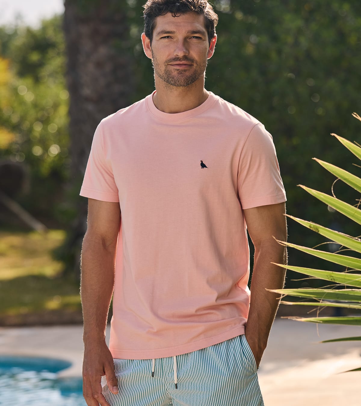 A man wearing a light pink crewneck t-shirt with a small navy ptarmigan logo on the left chest, paired with light blue and white striped swim trunks, standing outdoors near a pool with green foliage in the background.