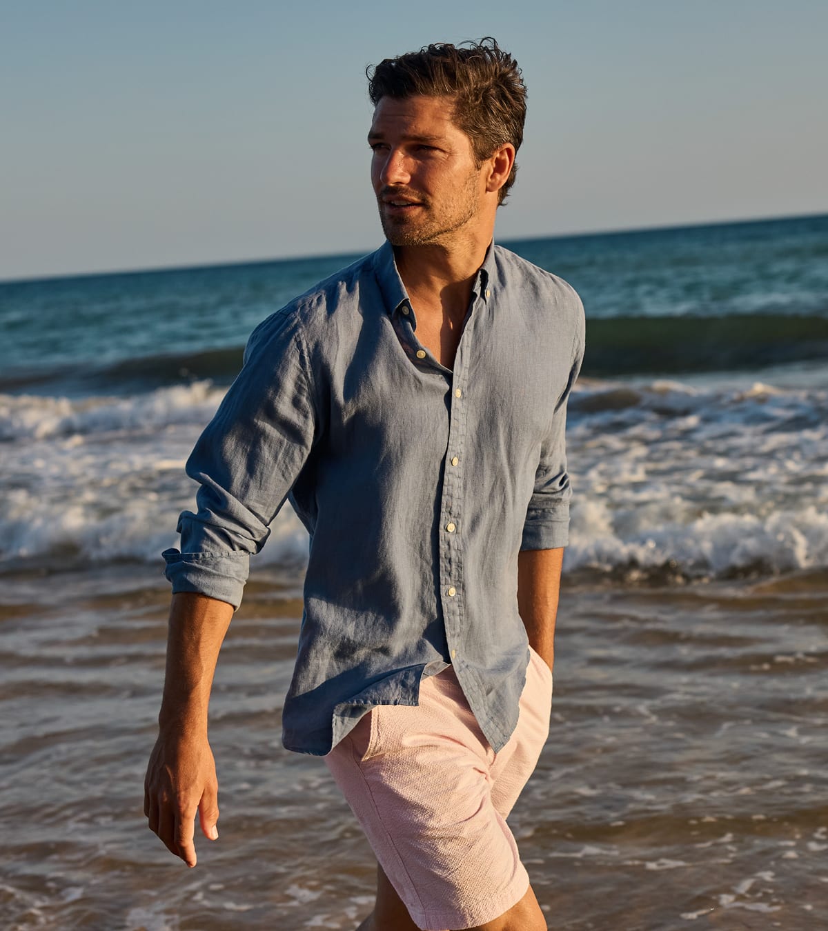 A man walking along a beach wearing a light blue linen button-down shirt and pale pink shorts.