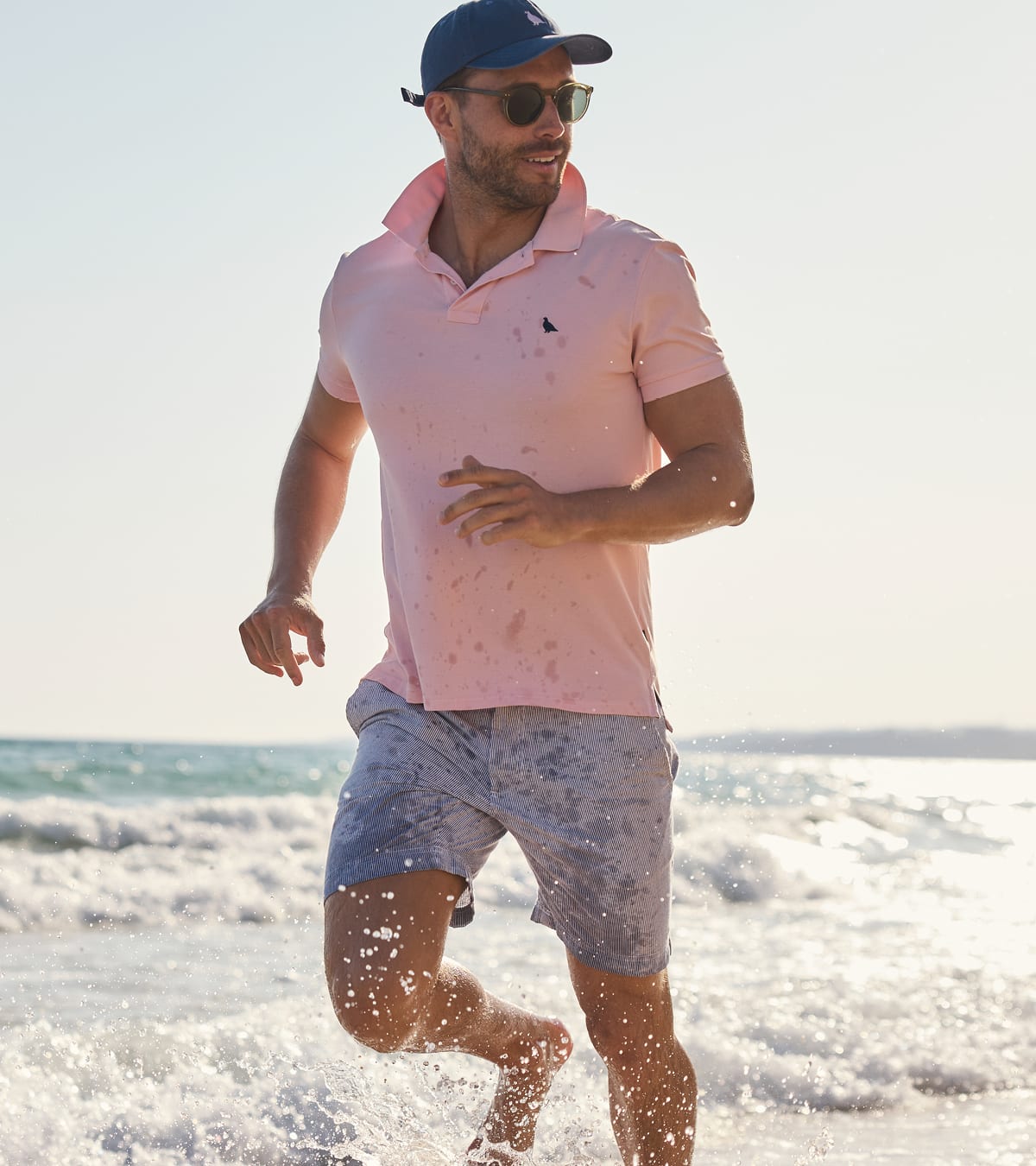 A man running through ocean waves on a beach wearing a light pink polo shirt with water splattered on it, matching water-damaged shorts, a navy cap, and sunglasses.
