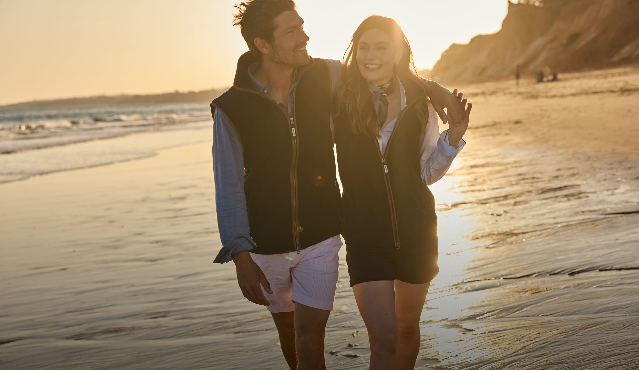 A couple walking arm in arm on a sandy beach during golden hour, wearing matching navy fleece gilets.