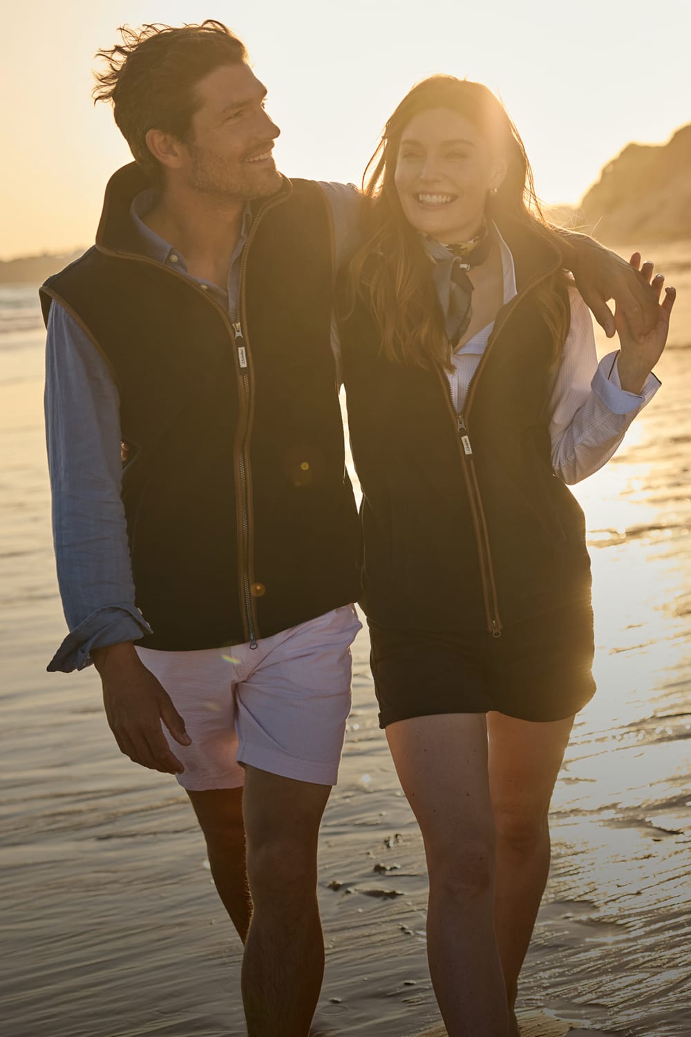 A smiling couple walking arm in arm on a beach at sunset. Both are wearing matching navy fleece gilets.