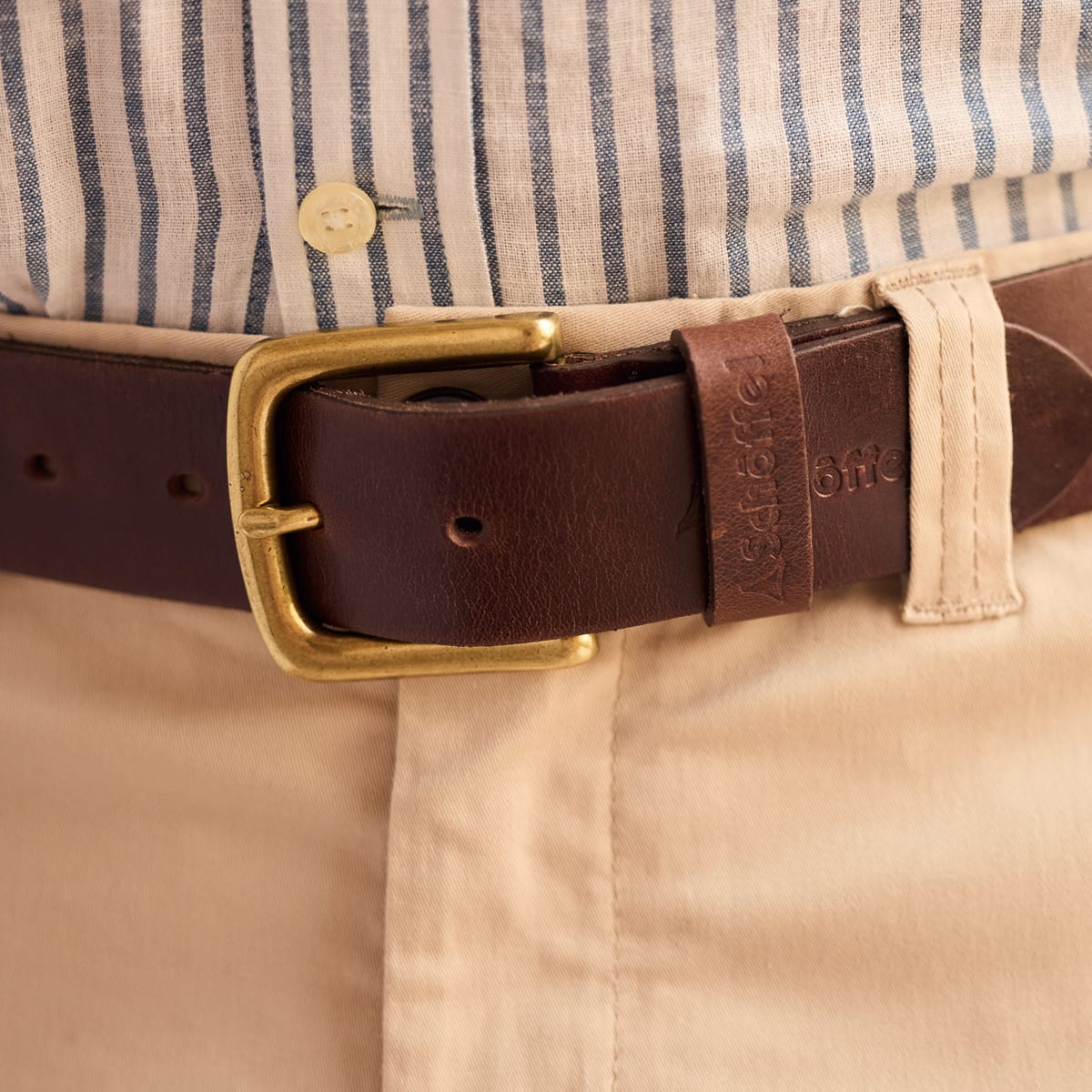 A close-up of a dark brown leather belt with a gold-tone buckle, worn at the waist over a beige shirt and khaki trousers.