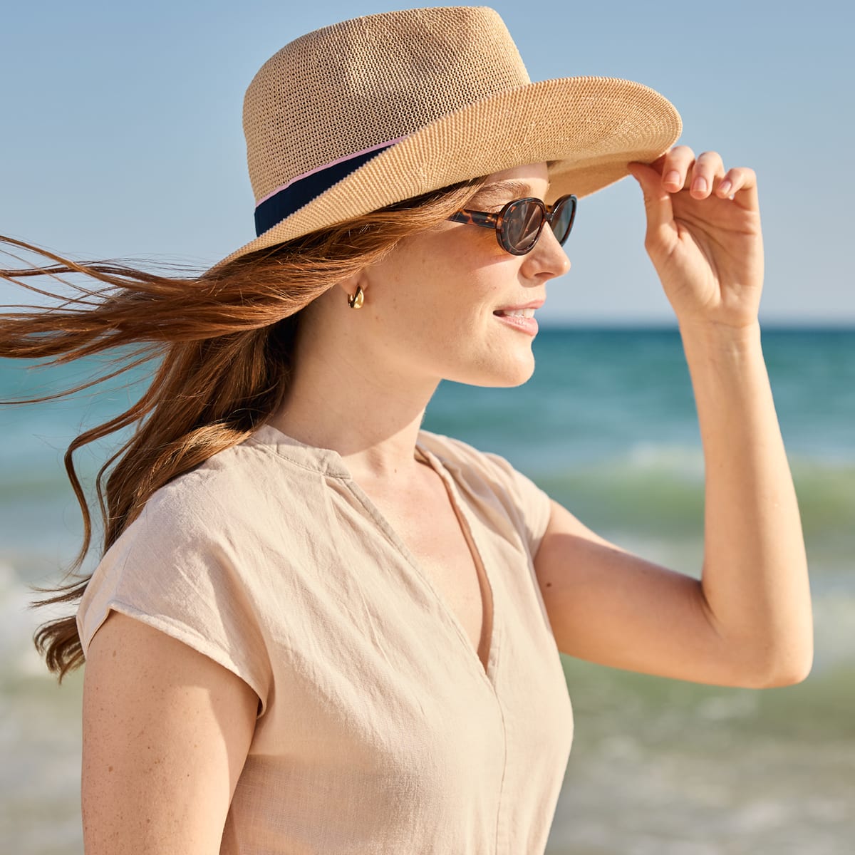 A woman wearing a beige woven straw sun hat with a navy band, sunglasses, and a light beige top, standing on a beach with the ocean and blue sky in the background.
