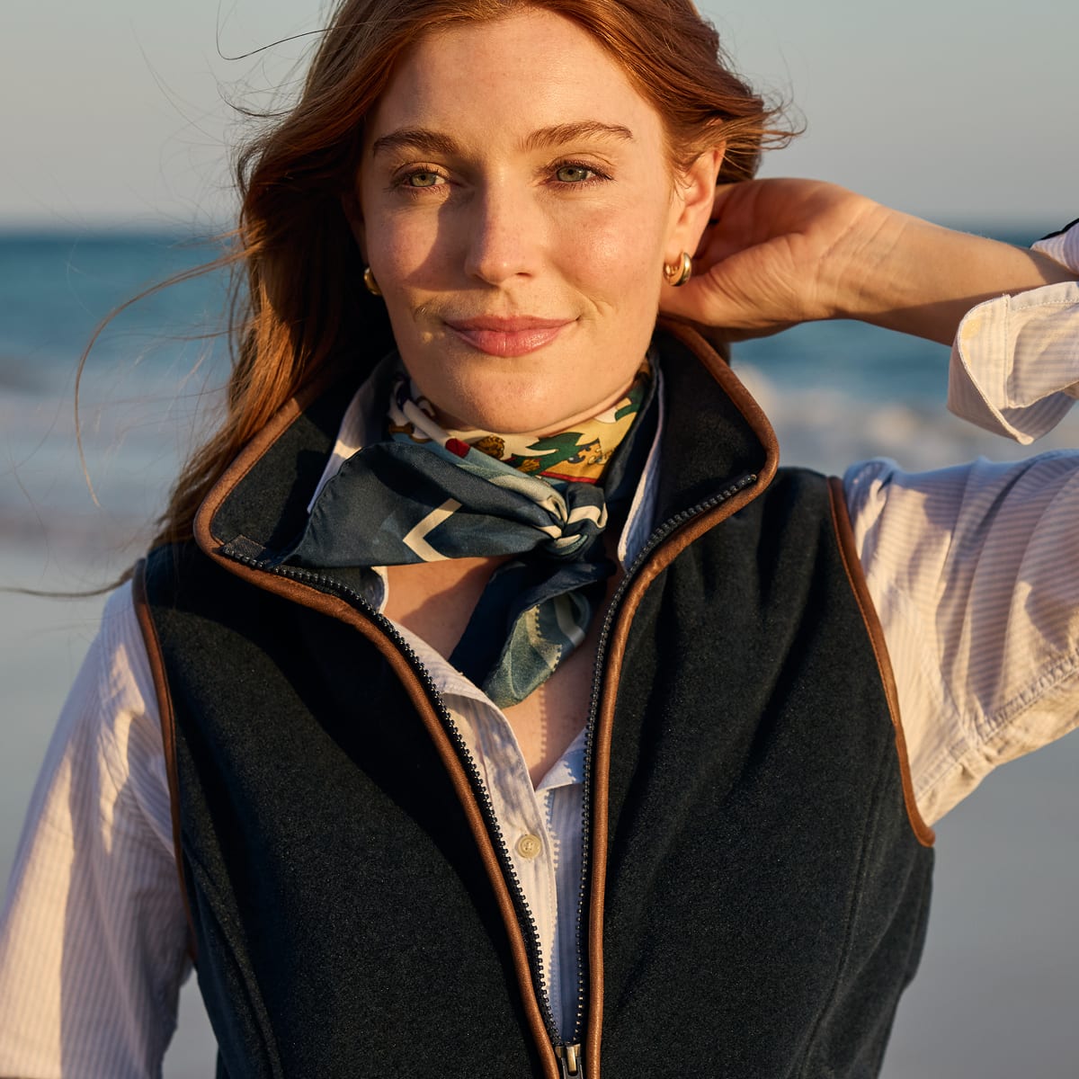 A woman on a beach wearing a navy fleece gilet  with brown trim over a white striped shirt and a patterned neck scarf.