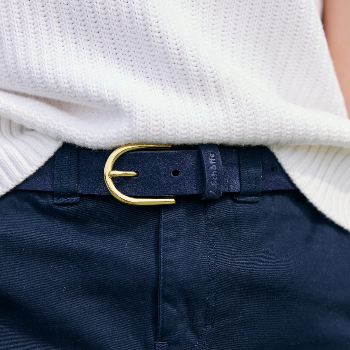A close-up of a person wearing a dark navy belt with a polished gold buckle, paired with dark navy trousers and a white knitted jumper.