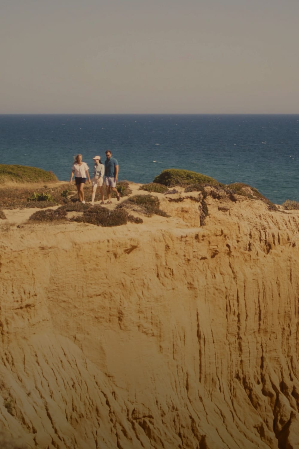 A scenic coastal landscape with a large sandy cliff in the foreground, blue ocean water in the distance, and three small people standing on the cliff edge under a pale sky.