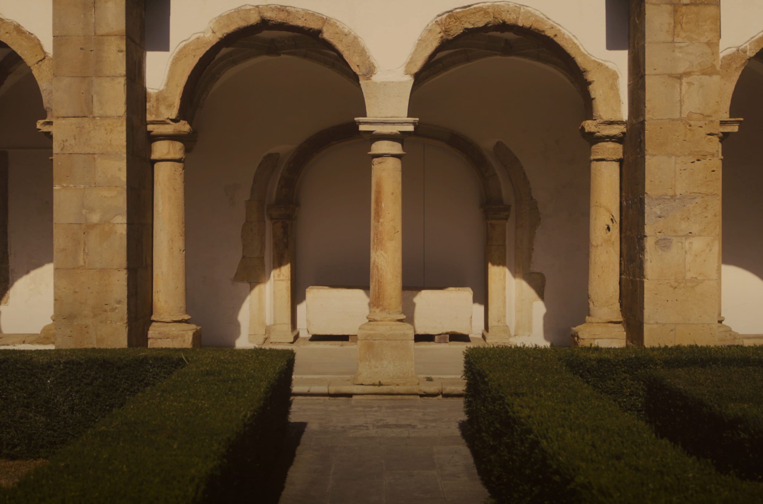 A sunlit stone cloister courtyard with arches and columns, leading toward a white bench in the center, with trimmed green hedges in the foreground.