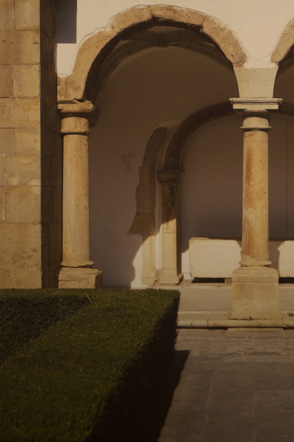 A warm-toned architectural photograph of a stone cloister with arches and columns, featuring a manicured green hedge in the foreground and a white bench