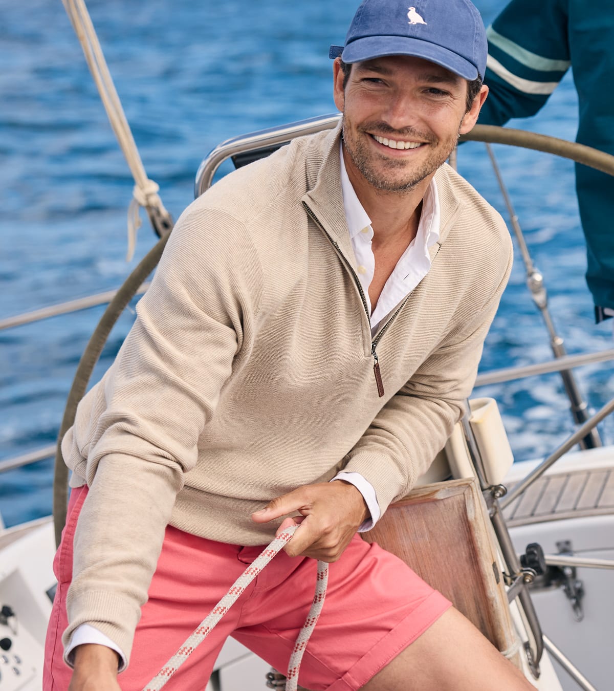A man on a sailboat wearing a beige quarter-zip jumper, coral pink shorts, a blue cap, and a white collared shirt, with blue water in the background.