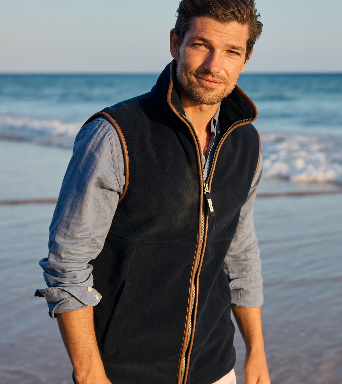 A man standing on a beach wearing a dark navy fleece zip-up gilet over a light blue long-sleeve shirt.