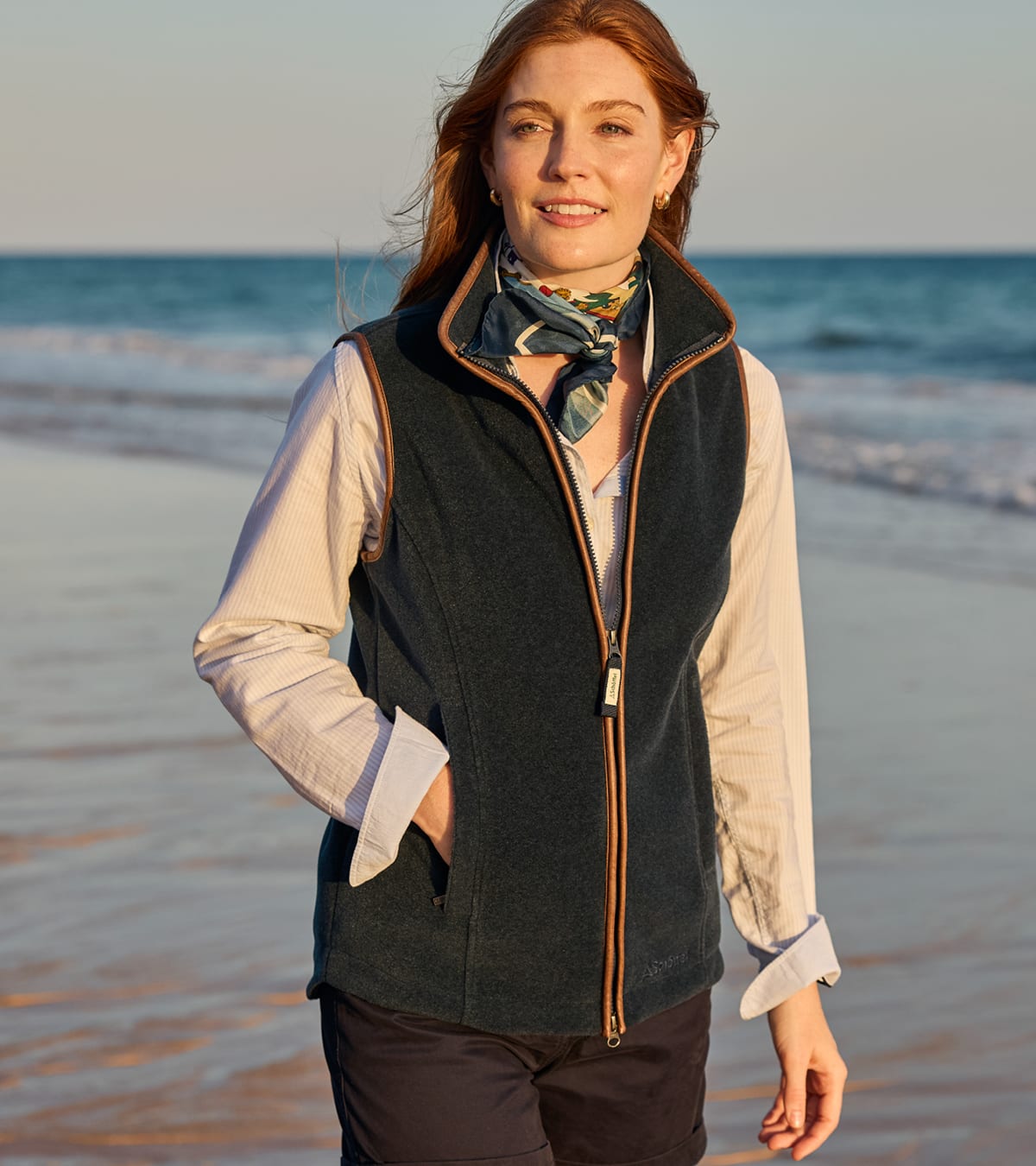 A woman standing on a beach wearing a dark navy fleece gilet over a light beige button-up shirt, accessorised with a patterned scarf.