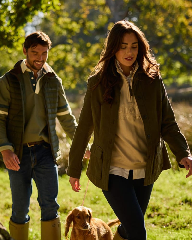 A man and a woman walking outdoors with a small dog on a grassy path. The woman wears an olive green coat over a beige pullover, and the man wears a green quilted jacket over a collared shirt and jeans.