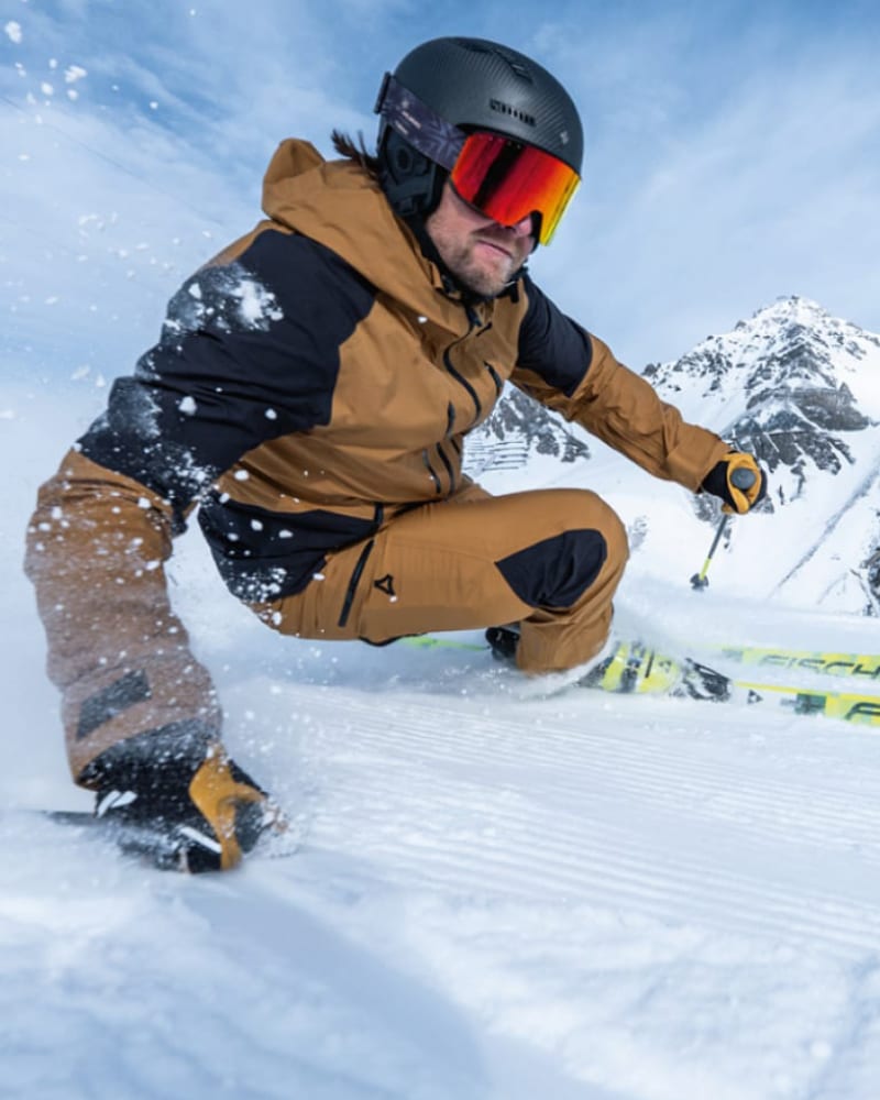 A skier in a crouched position carving through deep snow, wearing a tan and black ski suit, a dark helmet, and reflective orange-tinted goggles, with snowy mountains in the background.