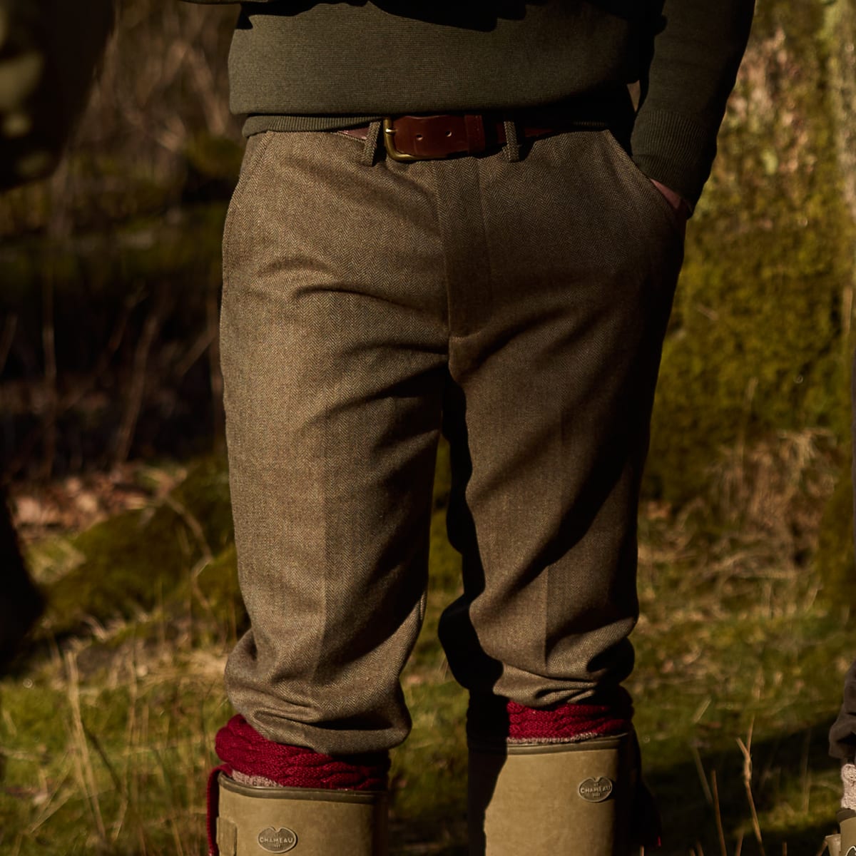 A person outdoors wearing olive green wool trousers, a dark green jumper, a brown leather belt, and tan rubber boots with red knit cuffs. The photo is cropped from the chest down, with warm natural light and a blurred forest background.