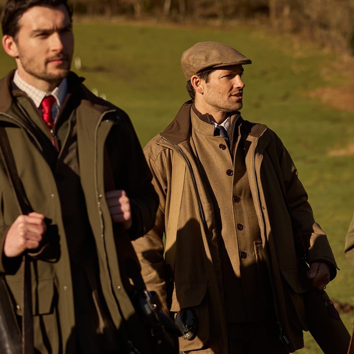 Two men outdoors in a grassy field wearing jackets and flat caps. The man on the right is more fully visible, wearing a brown tweed jacket, a light blue shirt, and a brown flat cap, while the man on the left is partially cropped and wears an olive green jacket.