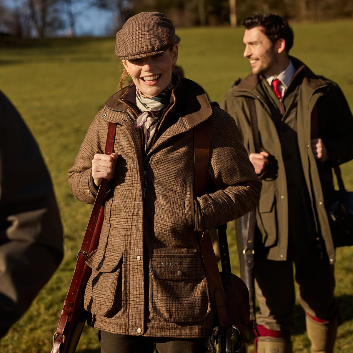 A woman in a brown plaid shooting jacket and flat cap smiles while holding a shotgun outdoors on a grassy field, with a man in a green shooting jacket standing behind her.