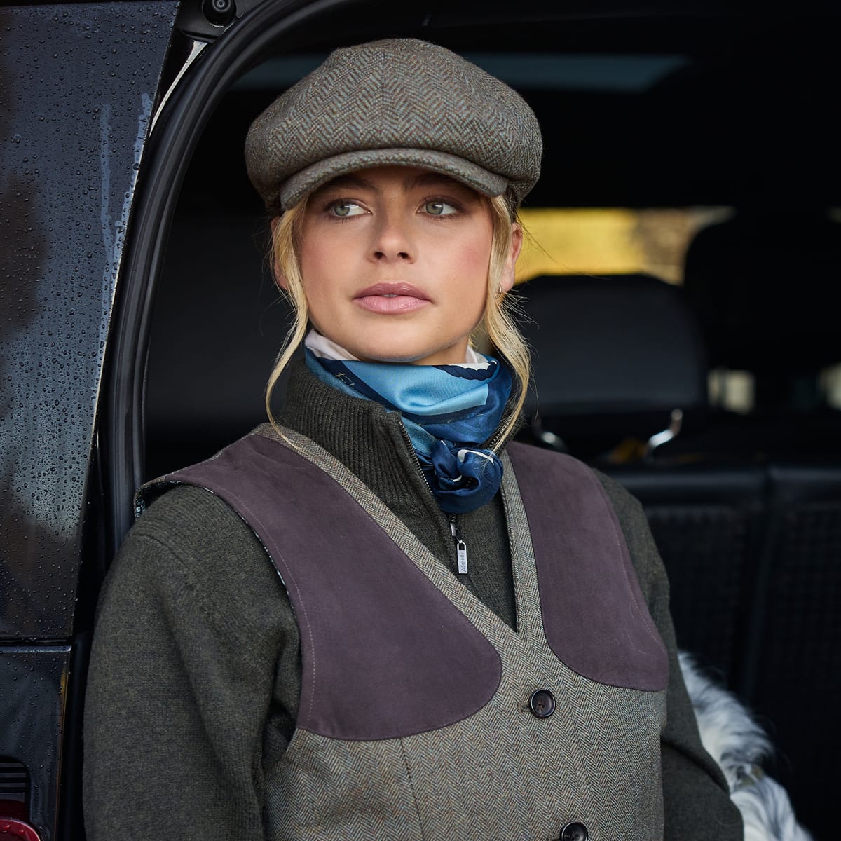 A woman wearing a tweed flat cap, a brown tweed waistcoat over a dark green jumper standing in the doorway of a dark car.
