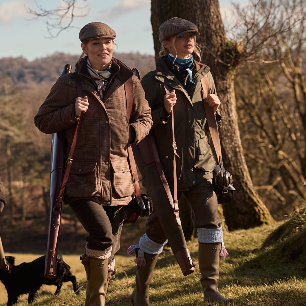 Two women walking outdoors in a wooded area, each carrying a shotgun over their shoulder. They are dressed in brown and olive green shooting jackets with brown trousers and tall wellington boots.