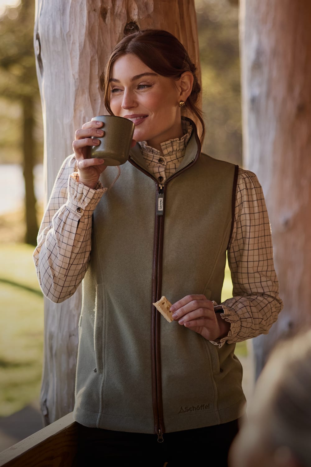 Woman wearing sage green zip-up fleece gilet over a beige checked shirt stands outdoors near a tree