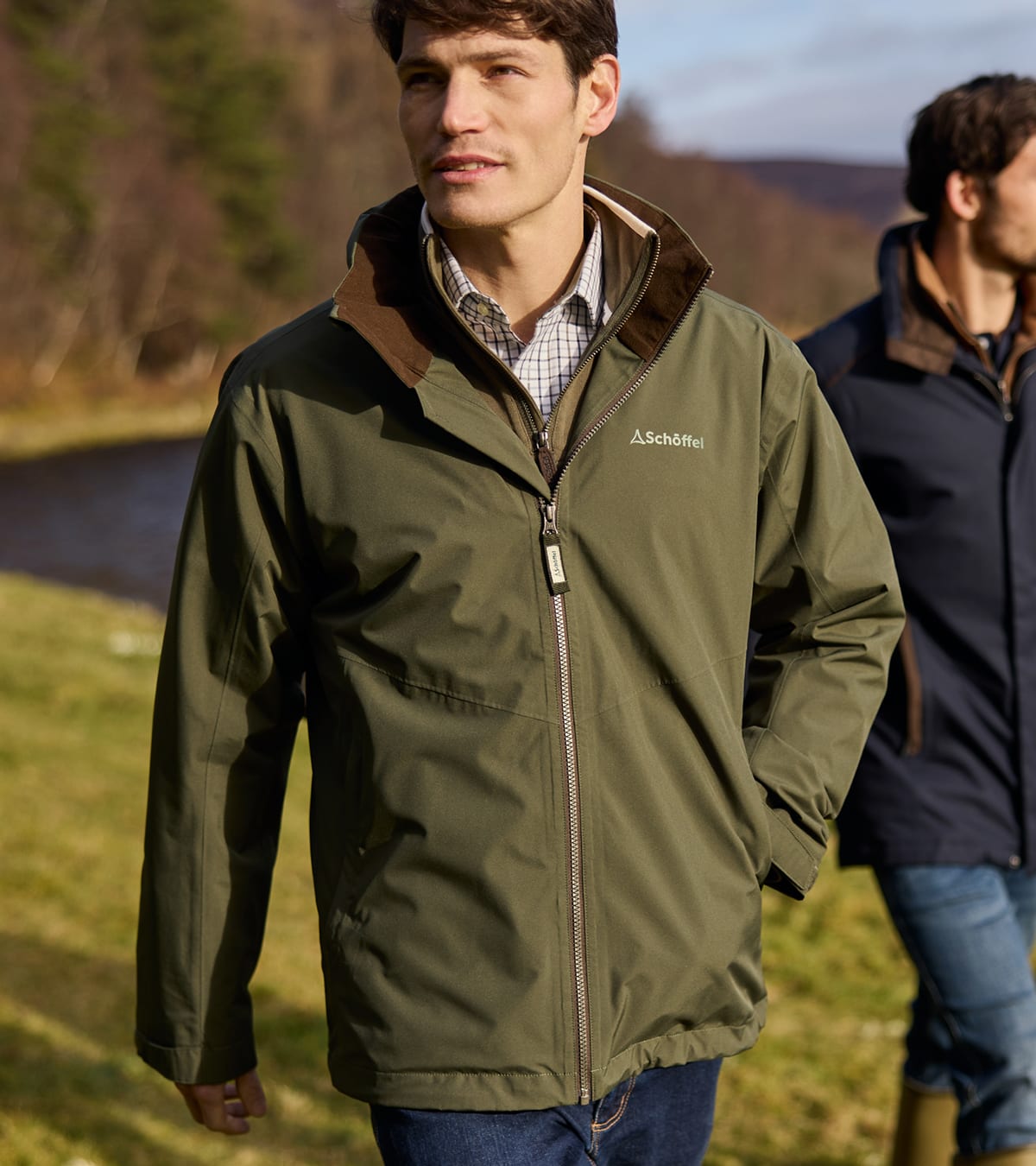 A man wearing an olive green jacket with brown collar trim, standing outdoors near a river with another man in the background.
