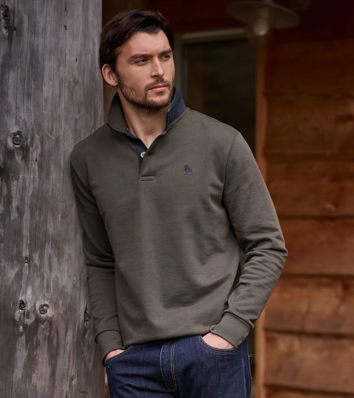 A man wearing a dark olive green long-sleeve sweatshirt with a blue inner collar, paired with blue jeans, leaning against a rustic wooden wall.