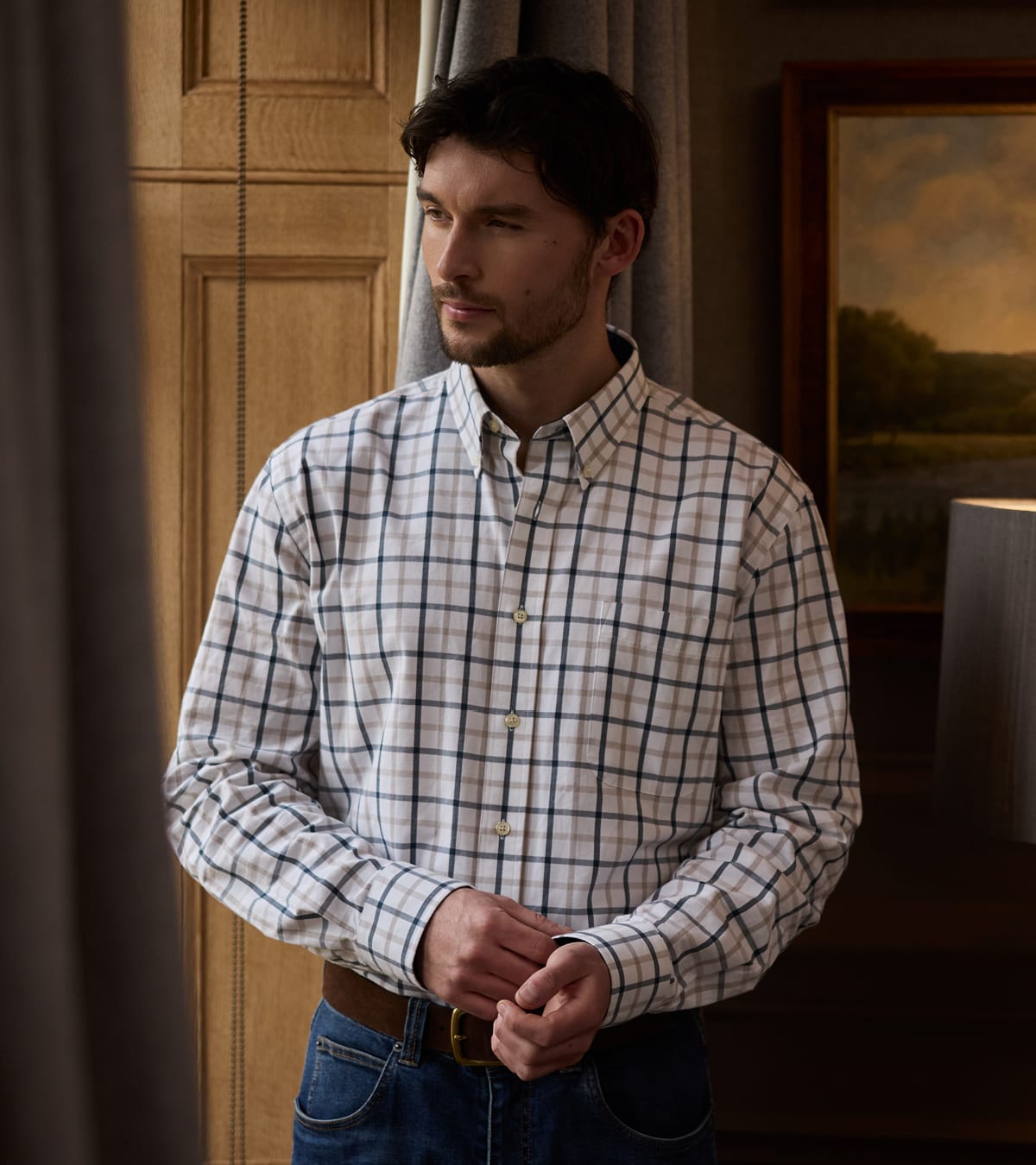 A man wearing a long-sleeve button-down shirt with a white, navy, and gray plaid pattern, paired with blue jeans and a brown leather belt, standing in a warmly lit room.