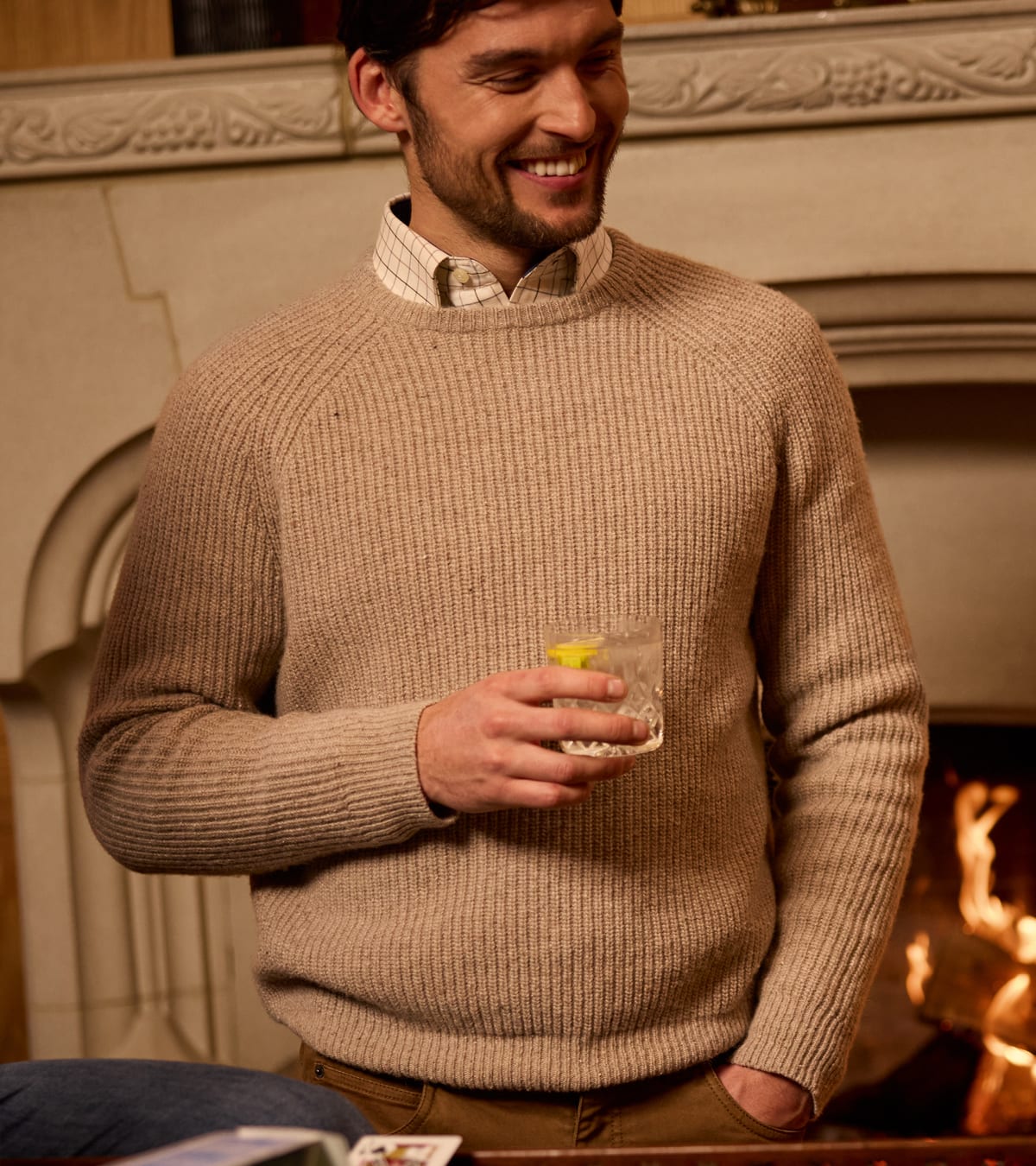 A man smiling in a cosy indoor setting, wearing a ribbed beige crewneck jumper over a collared shirt and holding a glass of water near a fireplace.