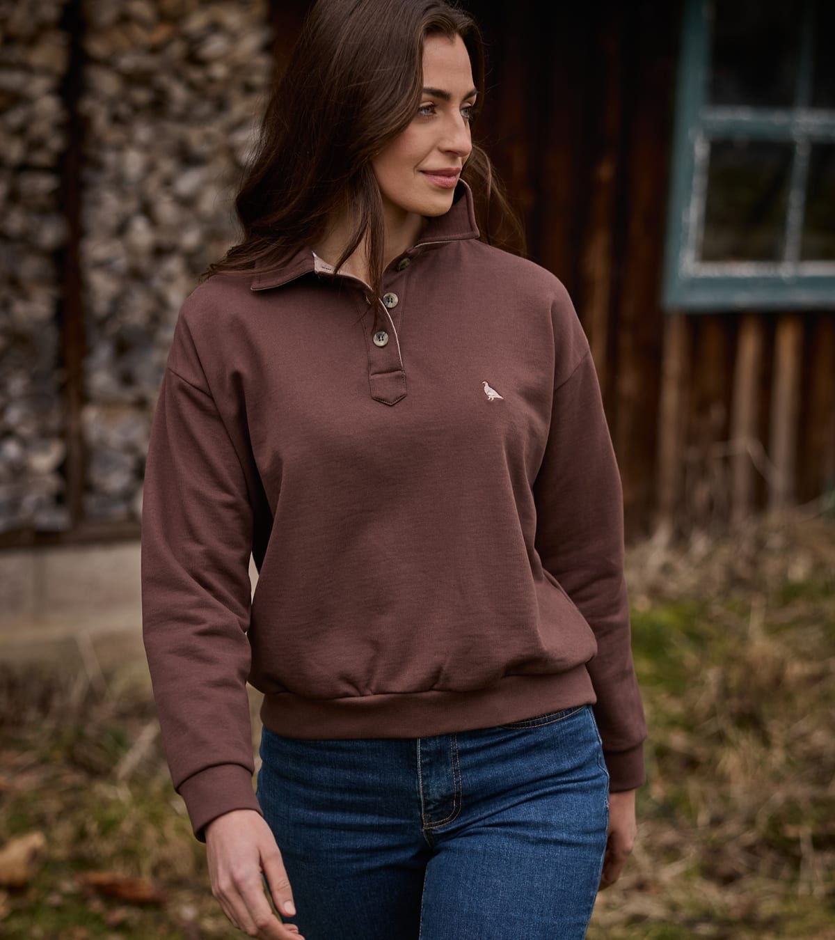 A woman wearing a brown long-sleeve quarter-placket pullover sweatshirt with a small embroidered  ptarmigan bird logo on the chest, paired with blue jeans, standing outdoors in front of a rustic wooden building.